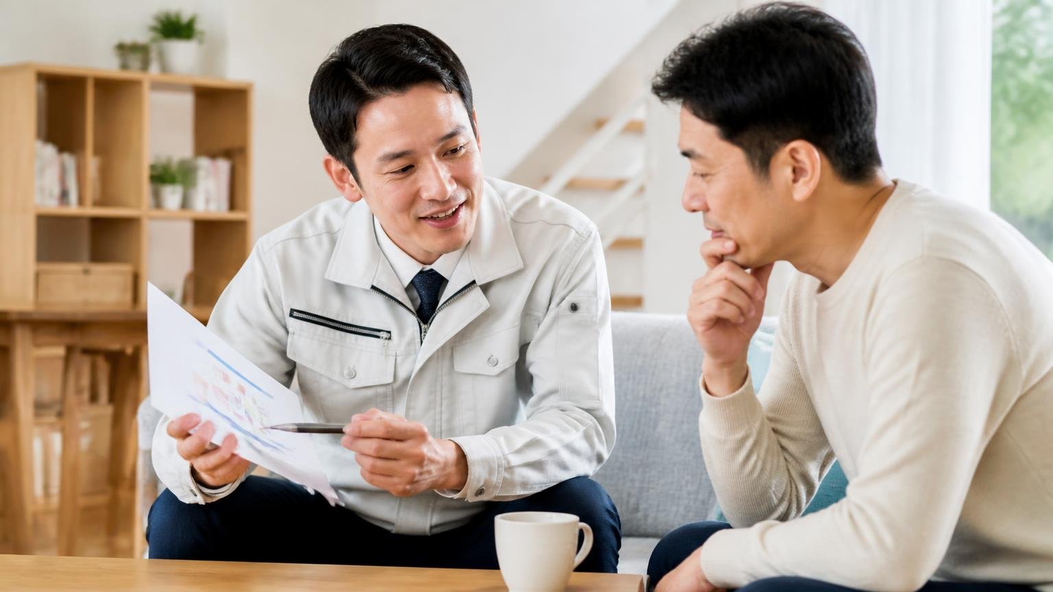 協会加盟の意味を説明する場面。住宅のリビングで作業着の男性1人が施主1人に書類を示して話している