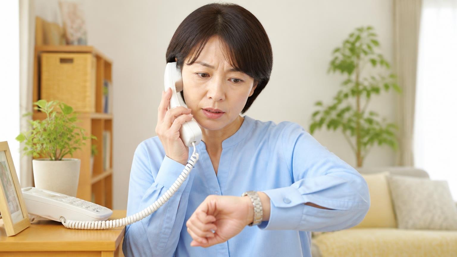 対応エリアと受付時間/自宅の固定電話の前で受話器を取る/40代女性1人/時計を見ながら電話をかける