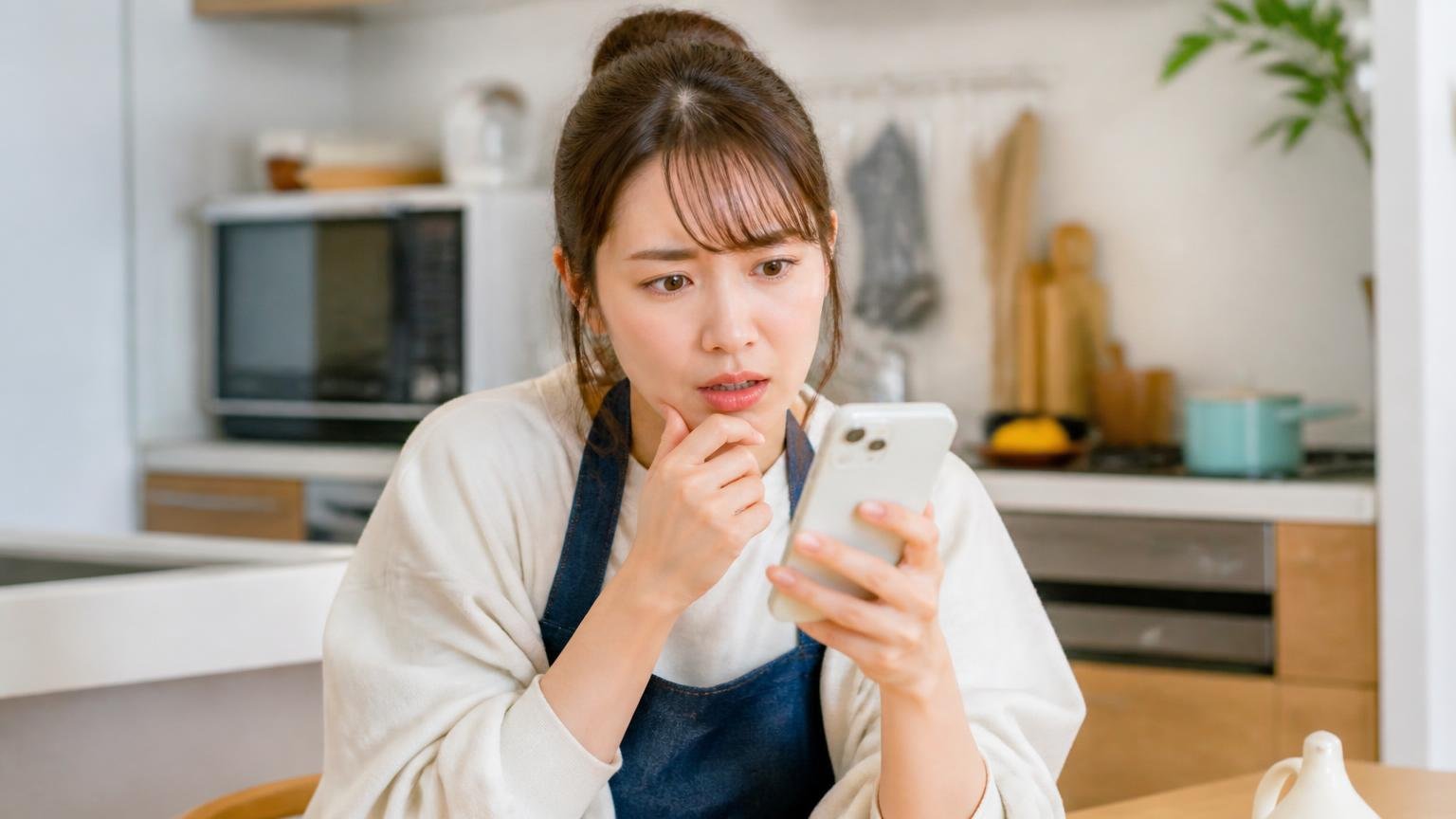 極端に安い表示価格をうのみにしない。台所でスマホの格安広告を見つめ困惑する20代女性
