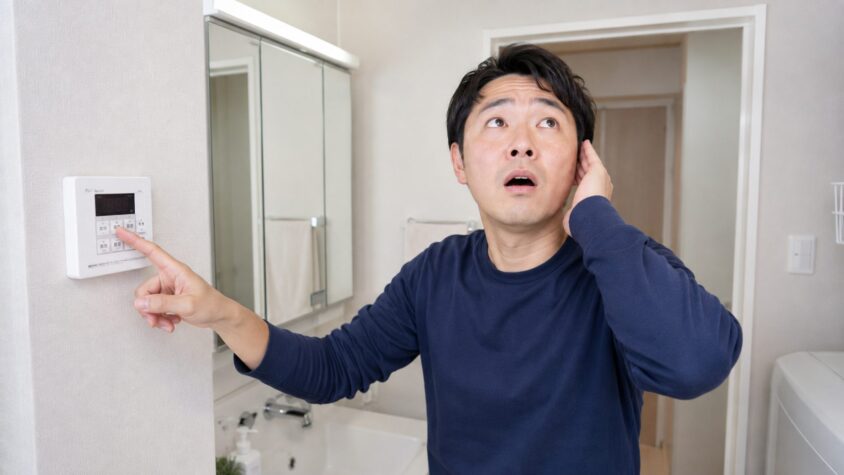給湯配管の膨張音。住宅の洗面所で給湯のリモコンを操作する30代男性1人が天井を気にする
