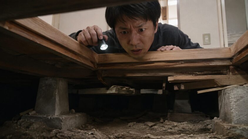 隙間だらけの床下を懐中電灯で照らす場面 賃貸住宅の床下点検口 30代男性が険しい表情で覗き込む
