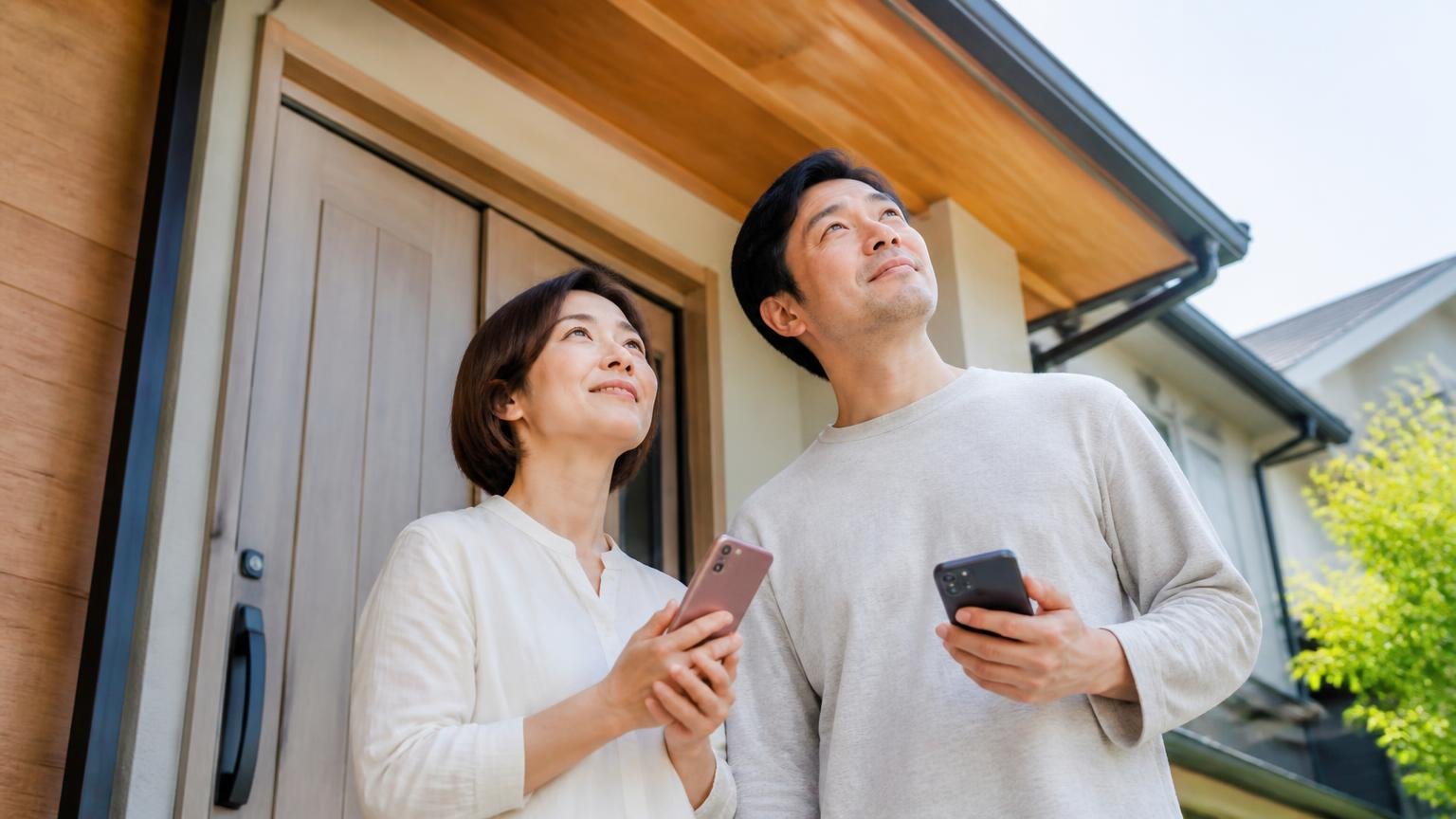 無料でできることから始めようと決める場面 木造戸建ての玄関前で軒下を見上げる夫婦2人 穏やかな表情でスマホを持つ