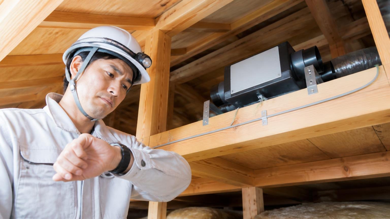 慣れはいつ起きるのか 木造戸建ての天井裏の梁の上に置かれた機器を見上げる作業員が腕時計を確認する