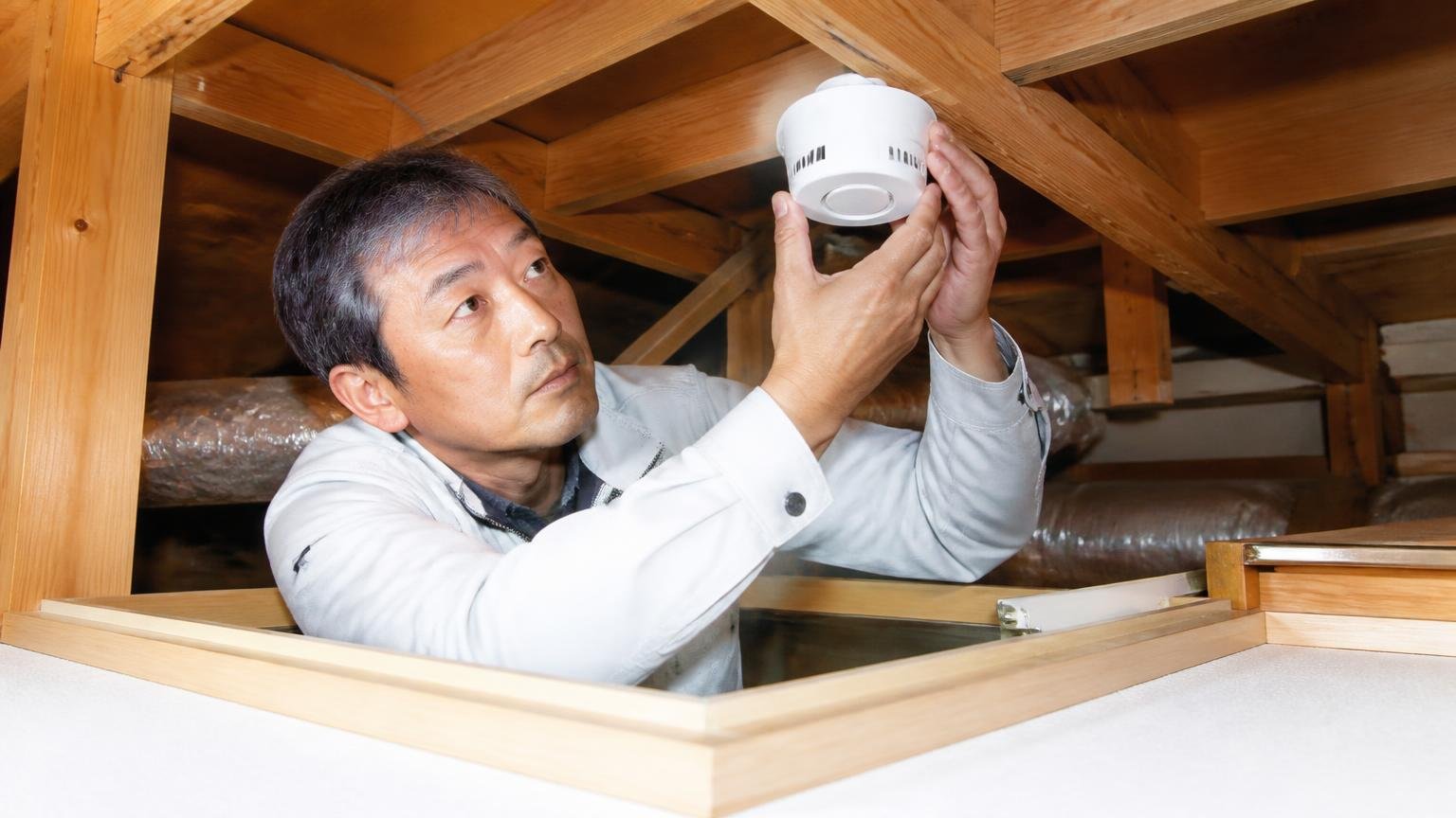設置場所と台数のコツ。木造戸建ての屋根裏の点検口から身を乗り出し機器を設置する50代男性